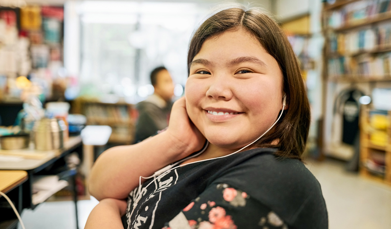 Uma mulher usando fones de ouvido olhando para a câmera e sorrindo. Ao fundo diversos livros em estantes