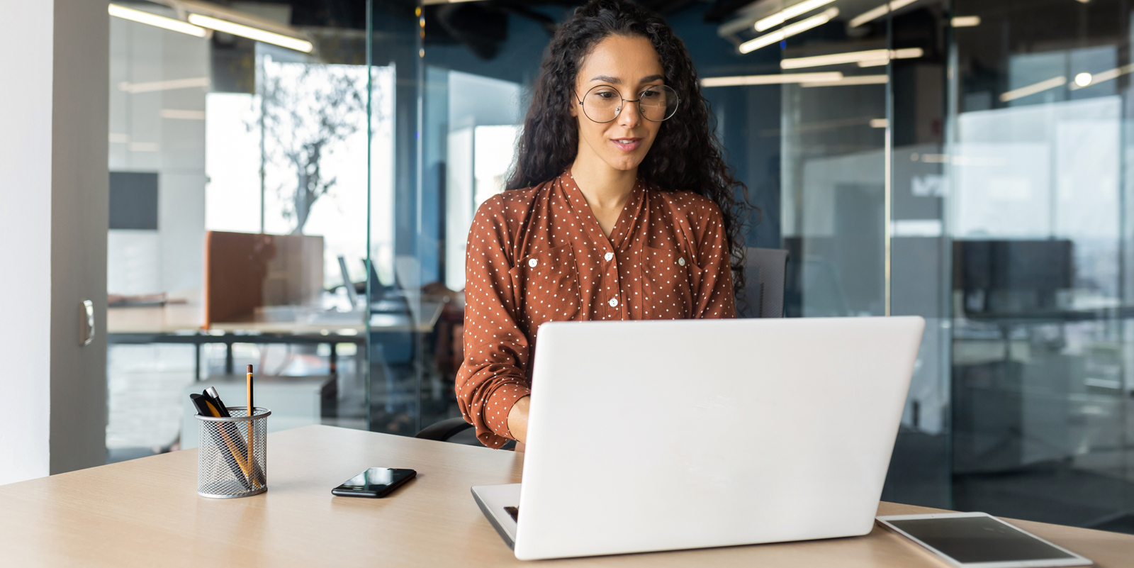 Uma pessoa trabalhando em um laptop em uma mesa de escritório moderno, com um smartphone e um porta-lápis por perto. Paredes de vidro e móveis de escritório ao fundo.