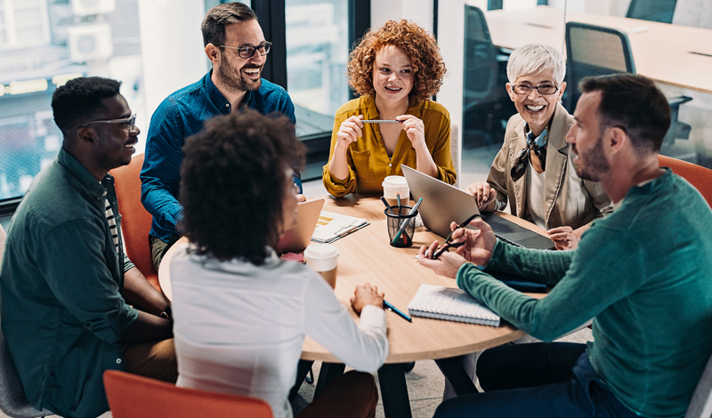 três homens e três mulheres sentados em volta de uma mesa de reunião com notebooks e cadernos para anotações