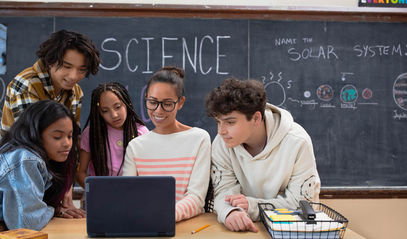 uma professora mostrando algo para os alunos em um notebook sobre a mesa. Ao fundo uma lousa com a palavra SCIENCE escrita