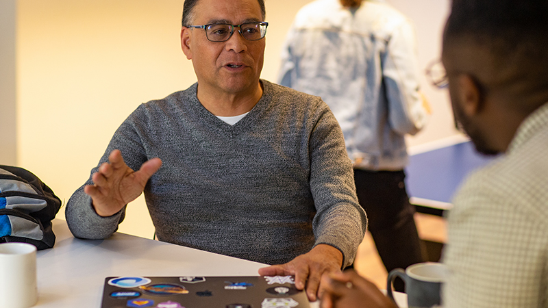 Duas pessoas conversando à mesa, com um laptop coberto de adesivos e uma caneca de café ao lado. Uma terceira pessoa é visível ao fundo.