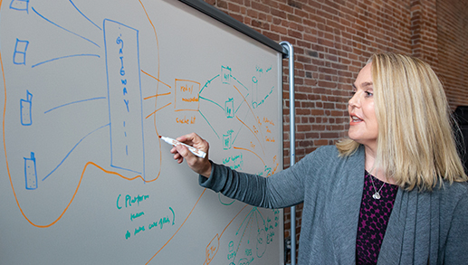Uma mulher desenha um diagrama técnico em um quadro branco em frente a uma parede de tijolos