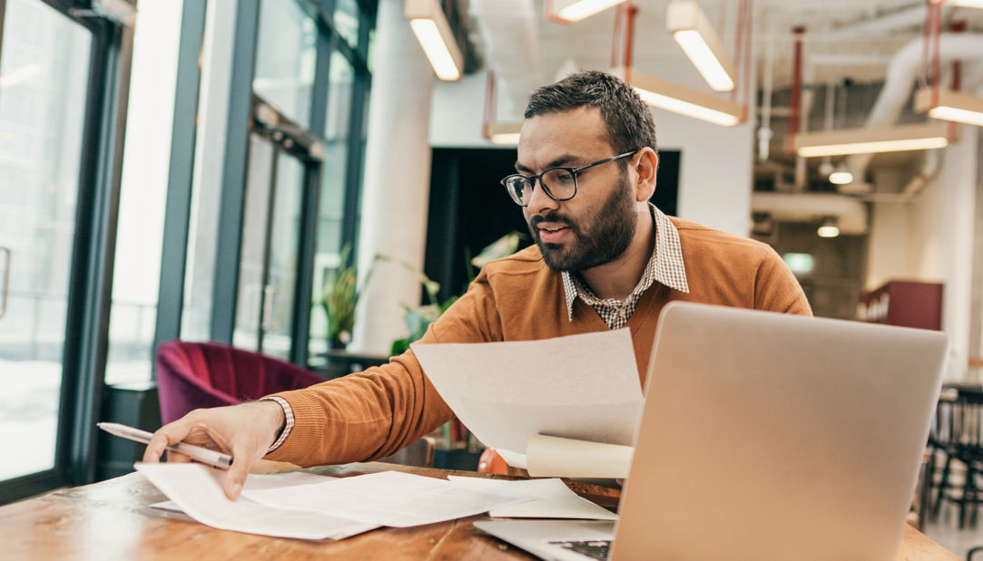 um homem sentado a mesa com diversos papéis e um notebook a frente