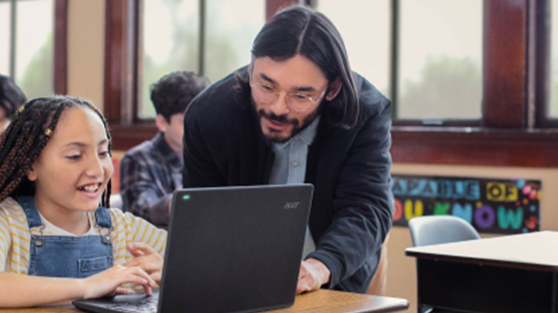 Um professor auxiliando um aluno que trabalha em um laptop em uma sala de aula, com outros alunos sentados ao fundo.