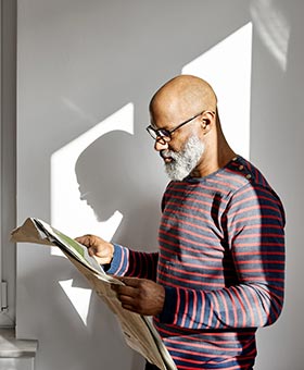 Homem lendo jornal em casa enquanto está em pé perto da janela.