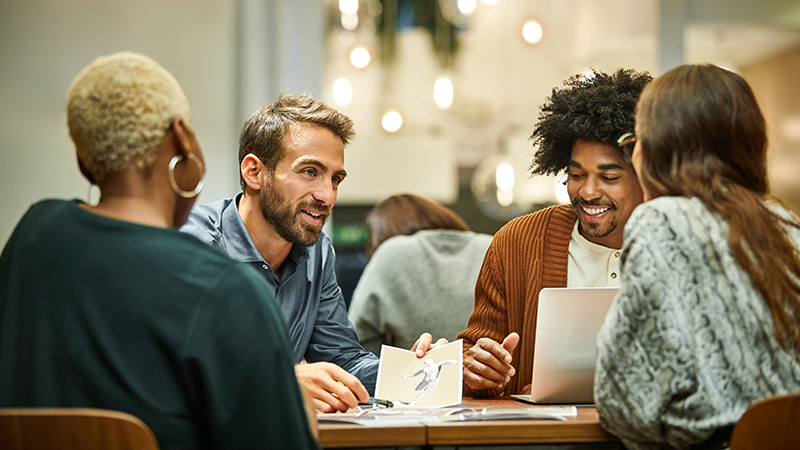 Um grupo de quatro pessoas senta-se ao redor de uma mesa, conversando. Duas delas estão olhando para um laptop, enquanto outra segura uma foto. Luzes suspensas e plantas são vistas ao fundo.