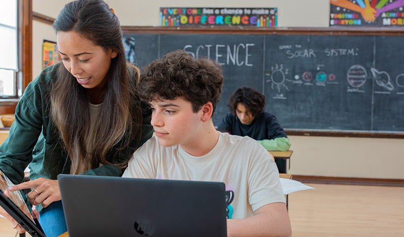 Professora ajudando um aluno com um tablet em uma sala de aula de ciências.