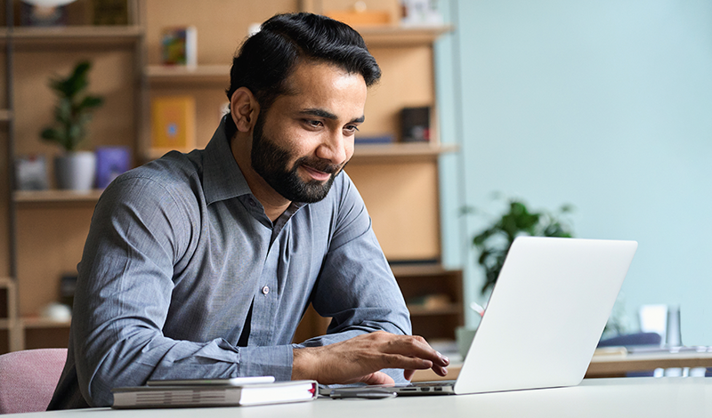 Um homem sorri enquanto trabalha em seu laptop em um escritório.