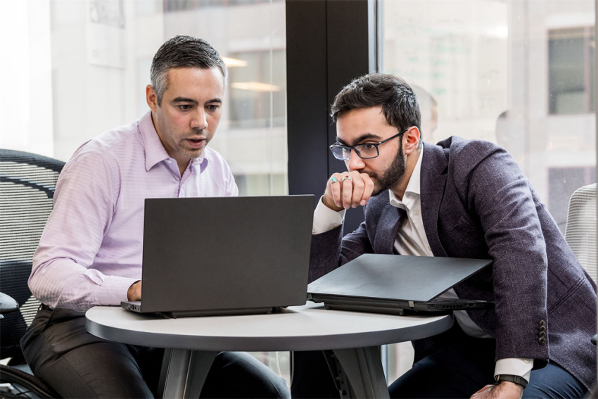 Two male office workers sitting at small round table in financial office, both looking at open laptop