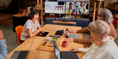 Tr&ecirc;s pessoas em uma mesa de reuni&atilde;o com o teams em um monitor