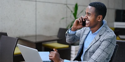 Homem negro sorrindo ao falar no celular com o computador &agrave; sua frente