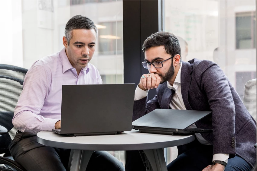 Two male office workers sitting at small round table in financial office, both looking at open laptop