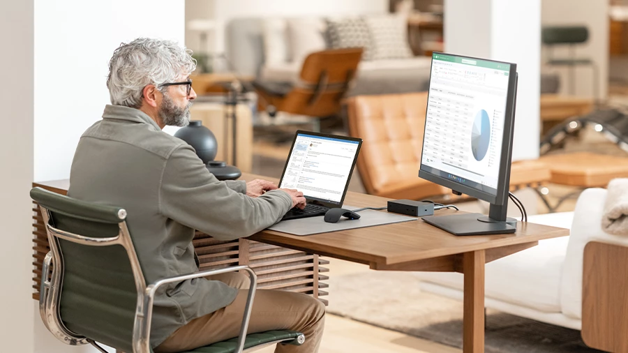 Homem sentado em frente ao computador, ele tem duas telas &agrave; sua frente. Em um est&aacute; sua caixa de entrada do Outlook e em outro, um gr&aacute;fico e uma tabela no Excel
